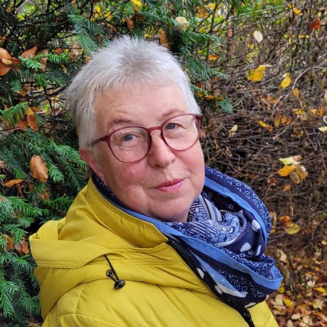 Older woman with short grey hair, red glasses, and yellow jacket against autumn foliage.