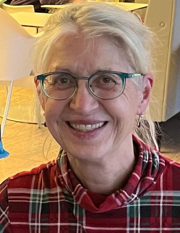 A smiling woman with grey hair, teal-framed glasses, and a red and green plaid shirt.